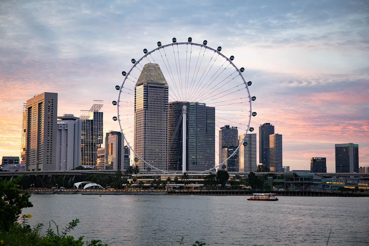 The Singapore Flyer observation wheel seen across the water at dusk, with city towers in front of and behind it and a pink sky above